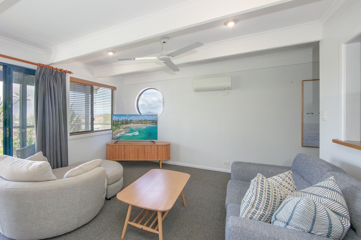 Ein modernes Wohnzimmer mit großem Fenster, Deckenventilator, Flachbildfernseher und Holzschrank.