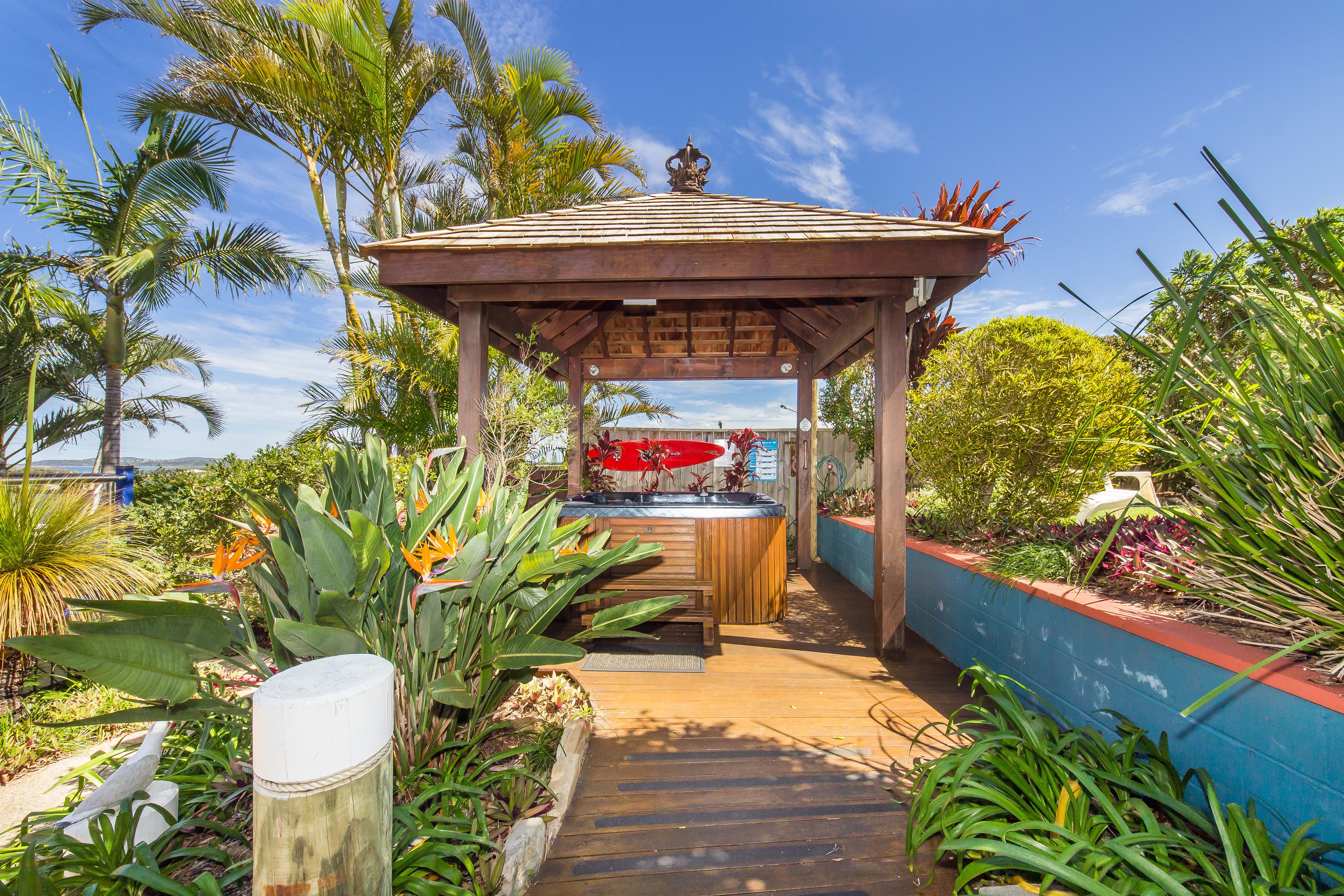 Um gazebo de madeira com hidromassagem, rodeado por plantas tropicais e um deck de madeira.