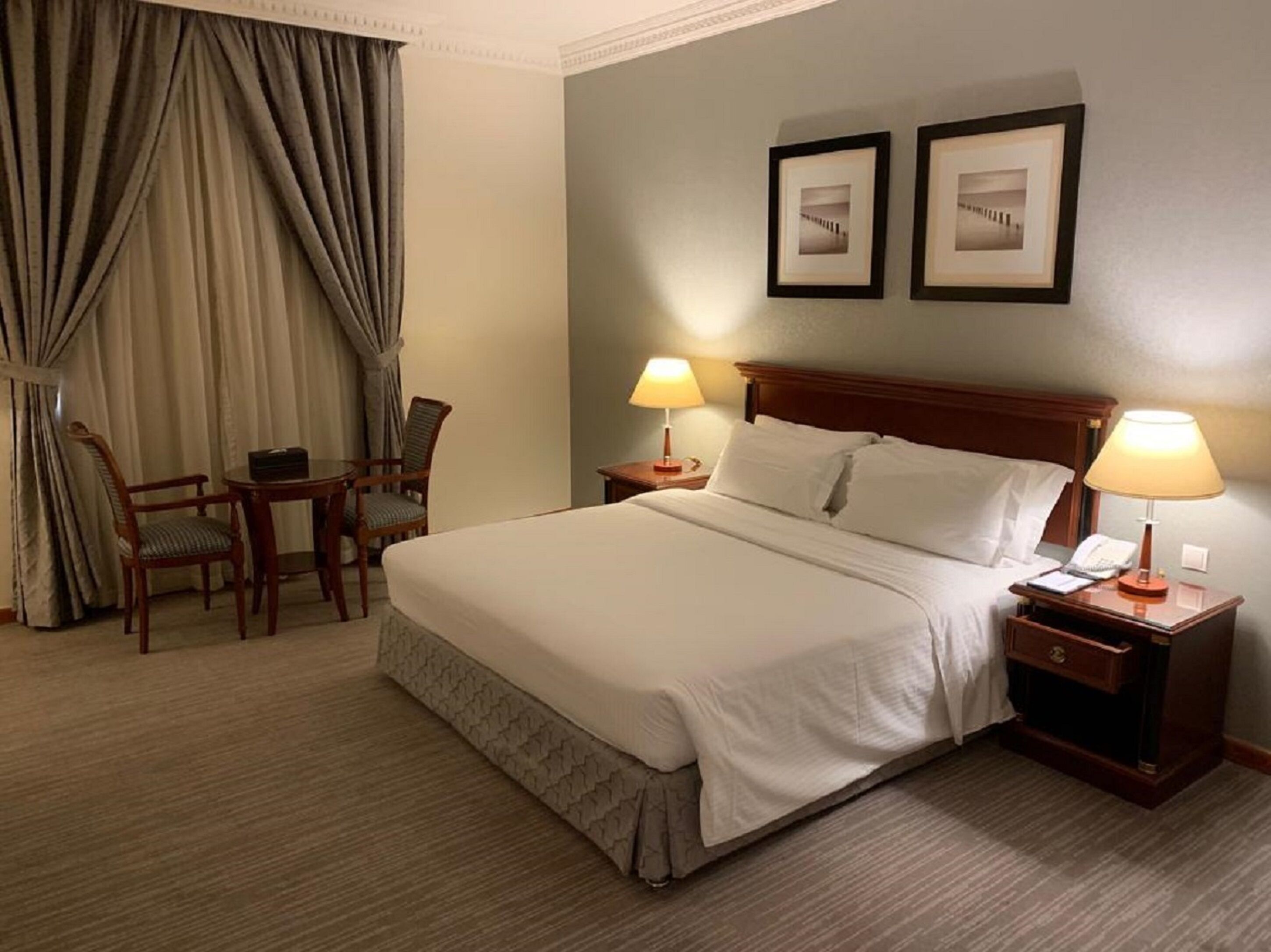 A hotel room with a large bed, two bedside tables with lamps, a chair, a desk, and two framed pictures on the wall.