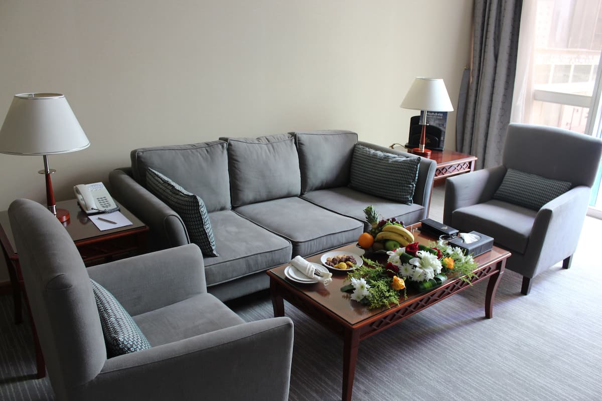 A modern hotel room with a grey sofa, a matching armchair, a wooden coffee table with fruit and flowers, and two bedside lamps.