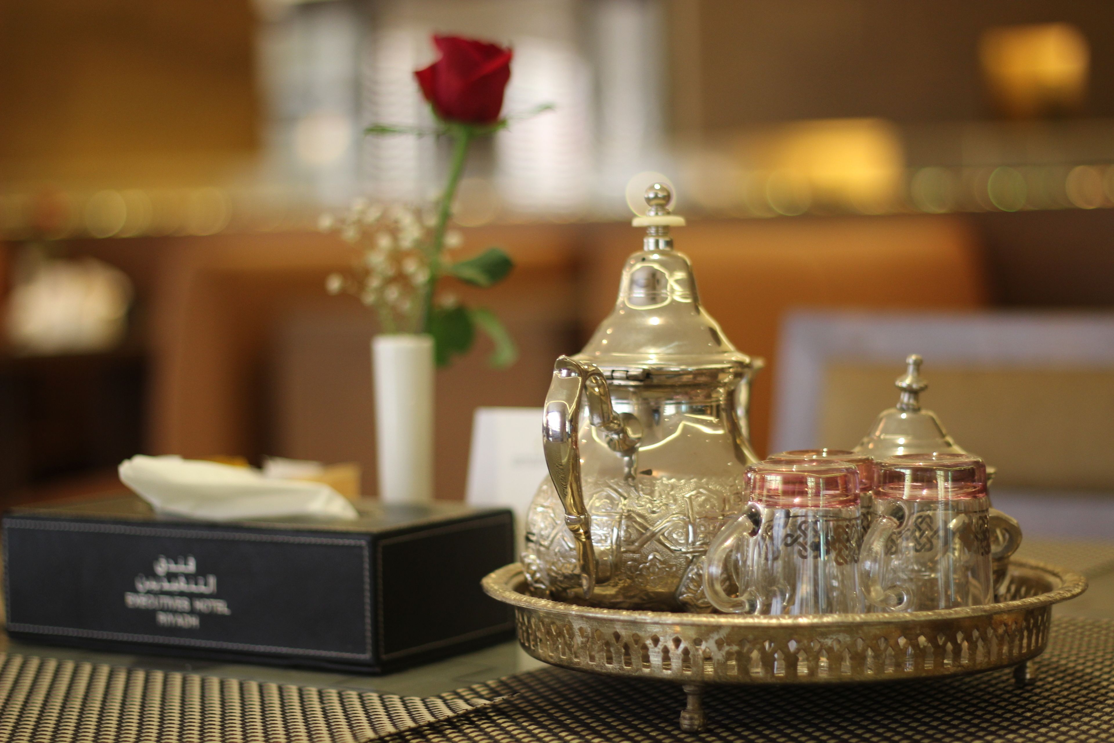 A tray with a teapot and sugar containers, a tissue box, and a rose in a vase on a table.