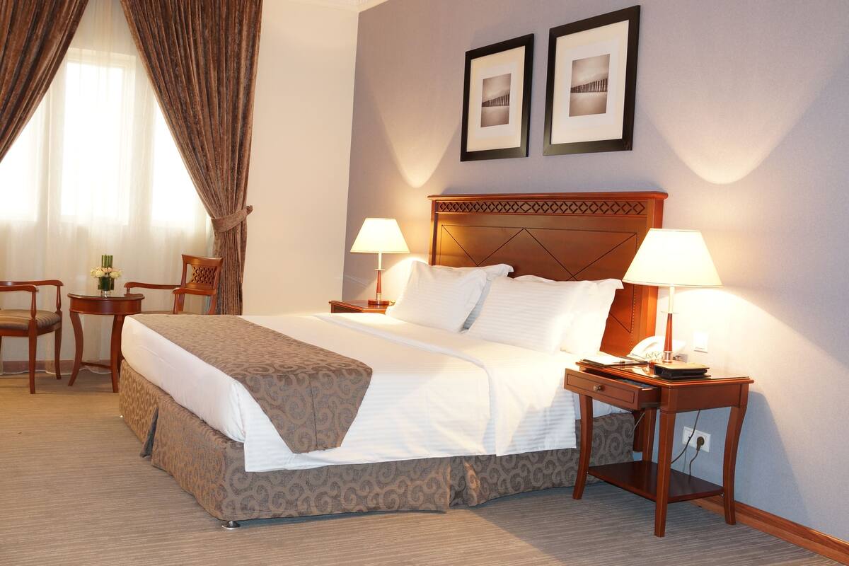 A hotel room with a large bed, two bedside tables with lamps, a chair, a small table with a vase, and two framed pictures on the wall.
