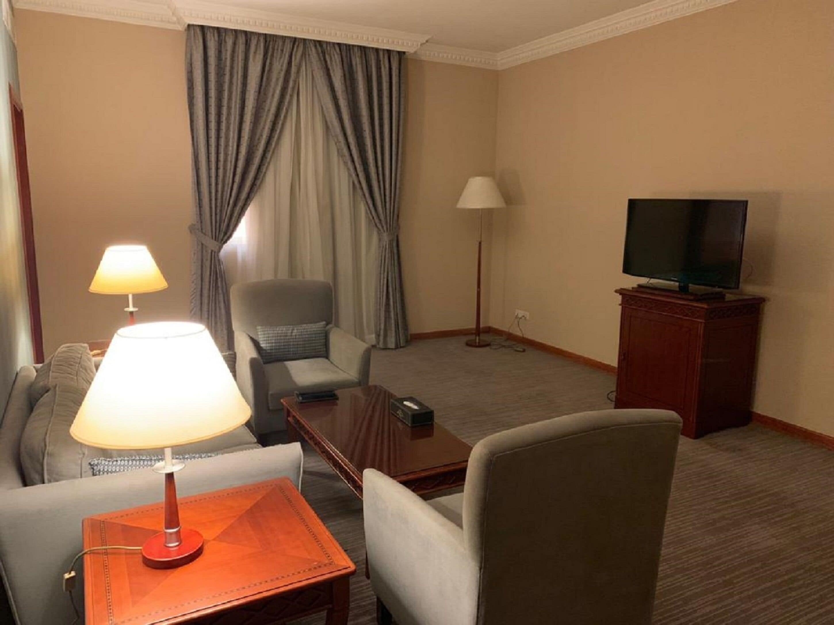 A hotel room with a sofa, armchair, a small wooden table with a lamp, a television on a cabinet, and a floor lamp.