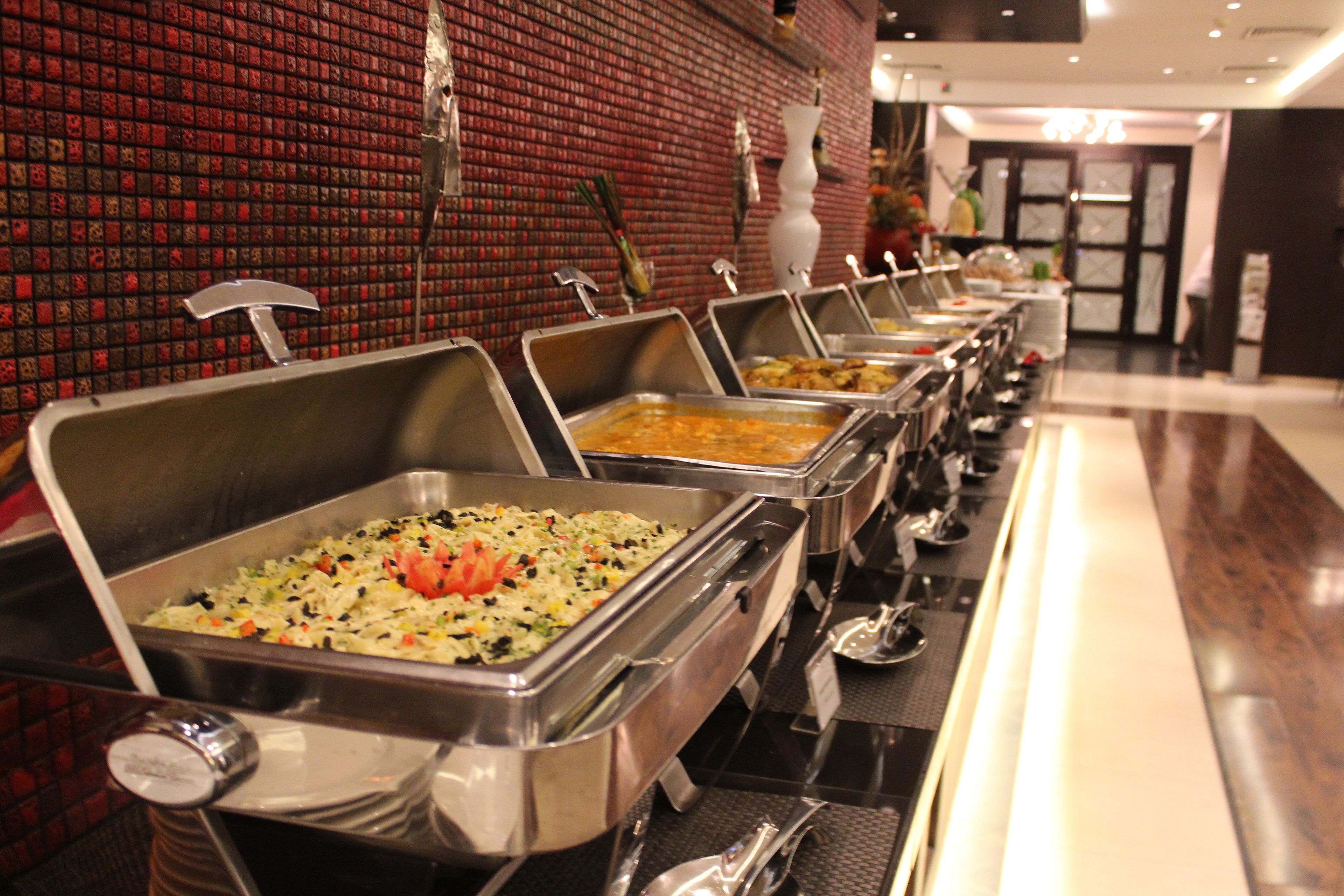 A buffet line with various dishes in metal serving trays, set against a red brick wall.