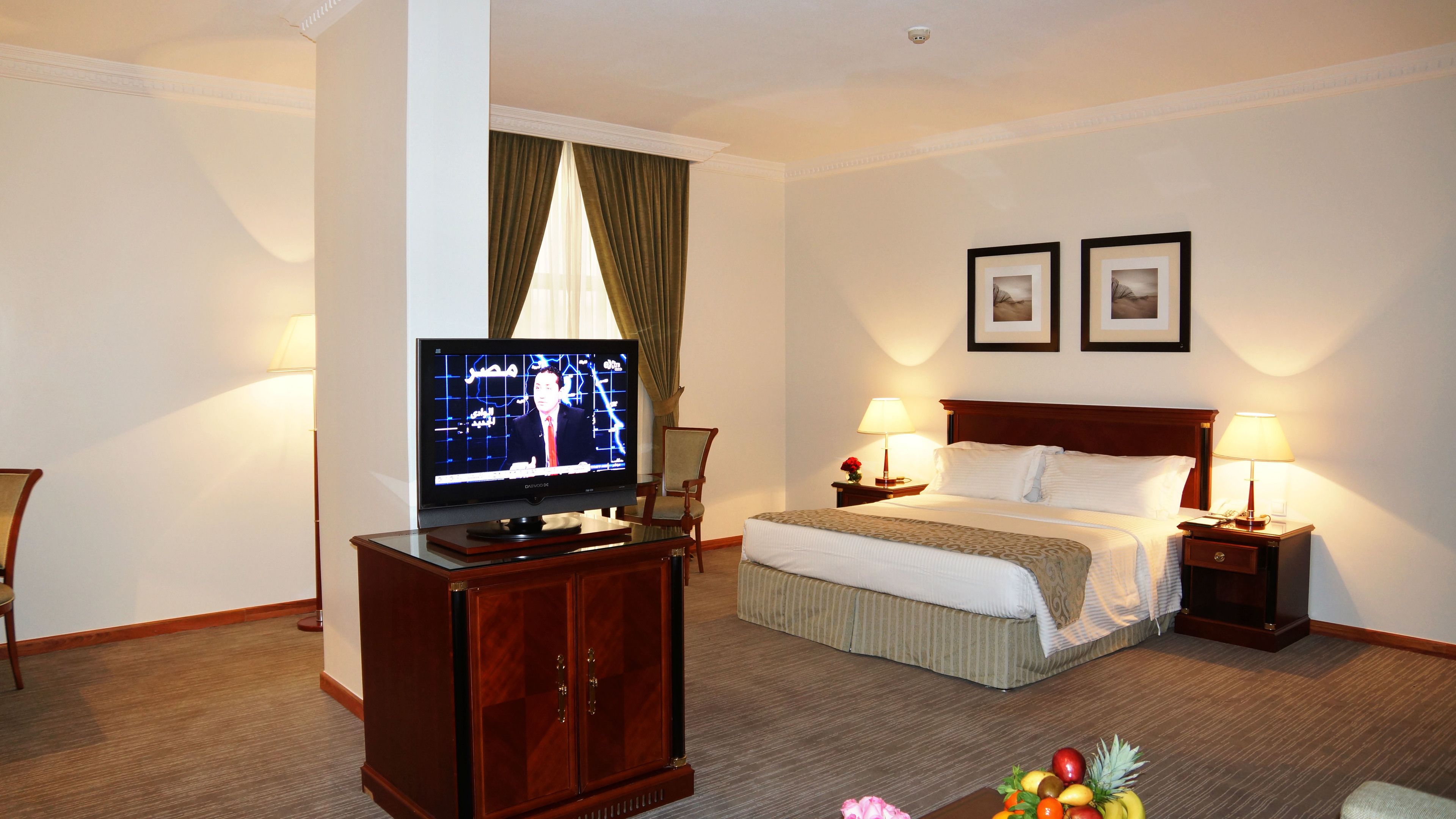Ein Hotelzimmer mit einem Bett, zwei Nachttischlampen, einem Fernseher auf einer Holzkommode, einem Sessel und einer Obstschale auf dem Boden.