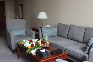 A hotel room with a grey sofa, a matching armchair, a wooden coffee table with a fruit bowl, a lamp, and a telephone.