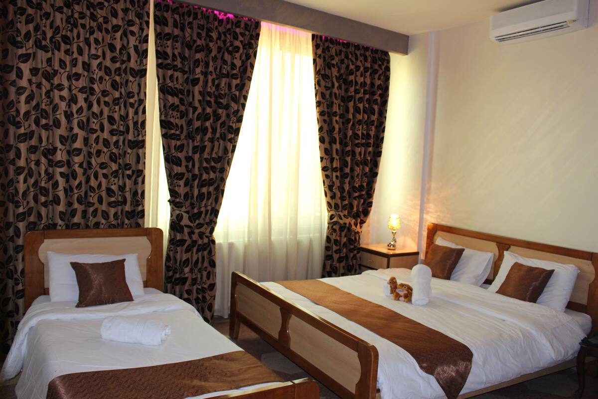 Une chambre d’hôtel avec deux lits, une tête de lit en bois, une table de chevet avec une lampe et des rideaux à motifs.
