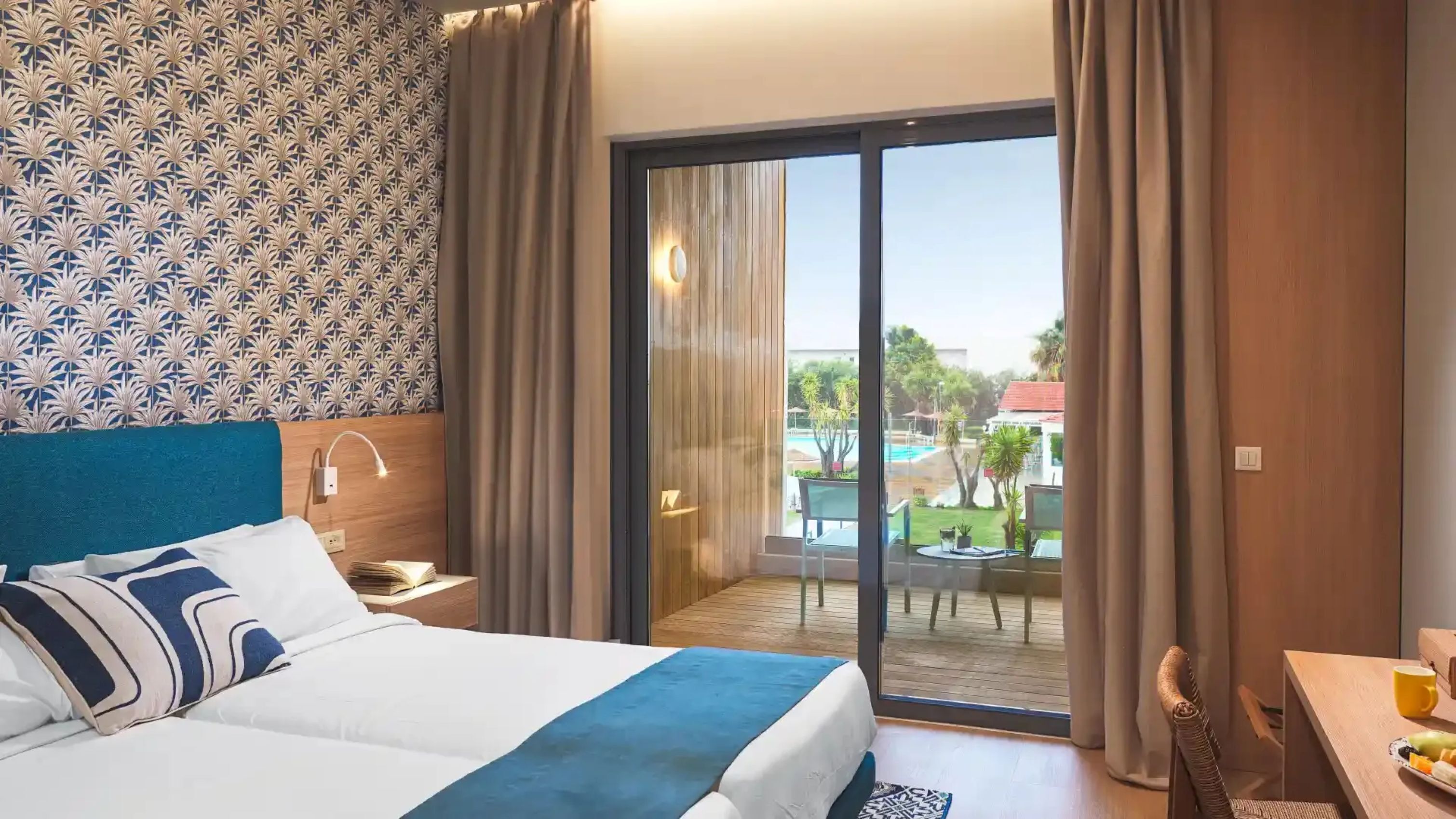 Habitación de hotel con cama, escritorio con silla, vista a una piscina y balcón con cortinas.