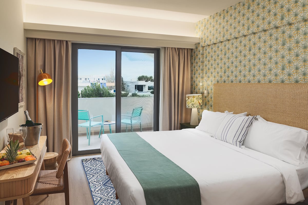 A hotel room with a bed, a desk with a bottle and glasses, a chair, and a view of the outside.