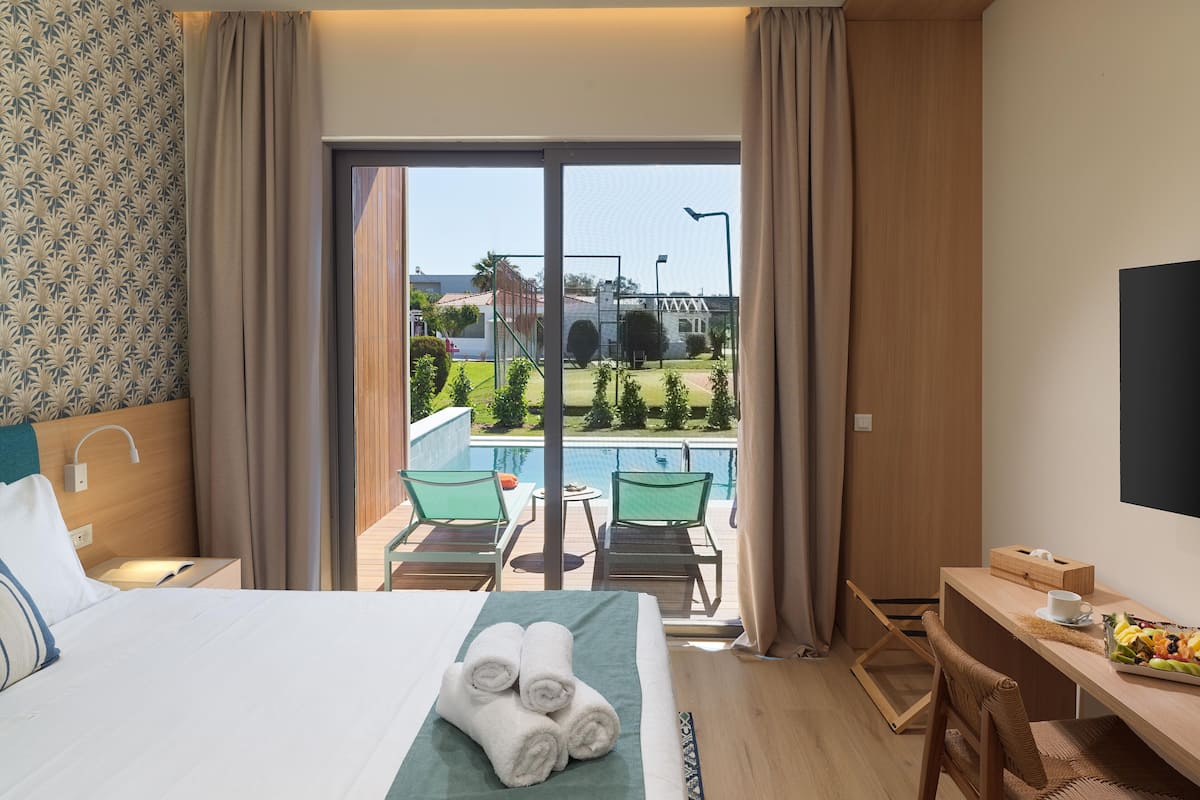 A hotel room with a bed, a desk, a chair, a table, and a view of a swimming pool and greenery.