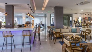 A modern hotel lobby with a bar area, seating, and a view of the kitchen in the background. - Leonardo Kolymbia Resort - All Inclusive (Rhodes)