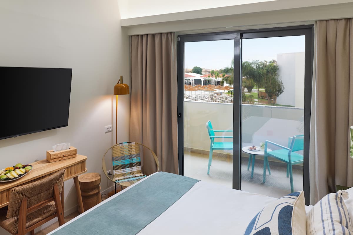 Habitación de hotel con cama, escritorio con ordenador portátil, televisor, balcón con mesa y sillas, y vistas al exterior.