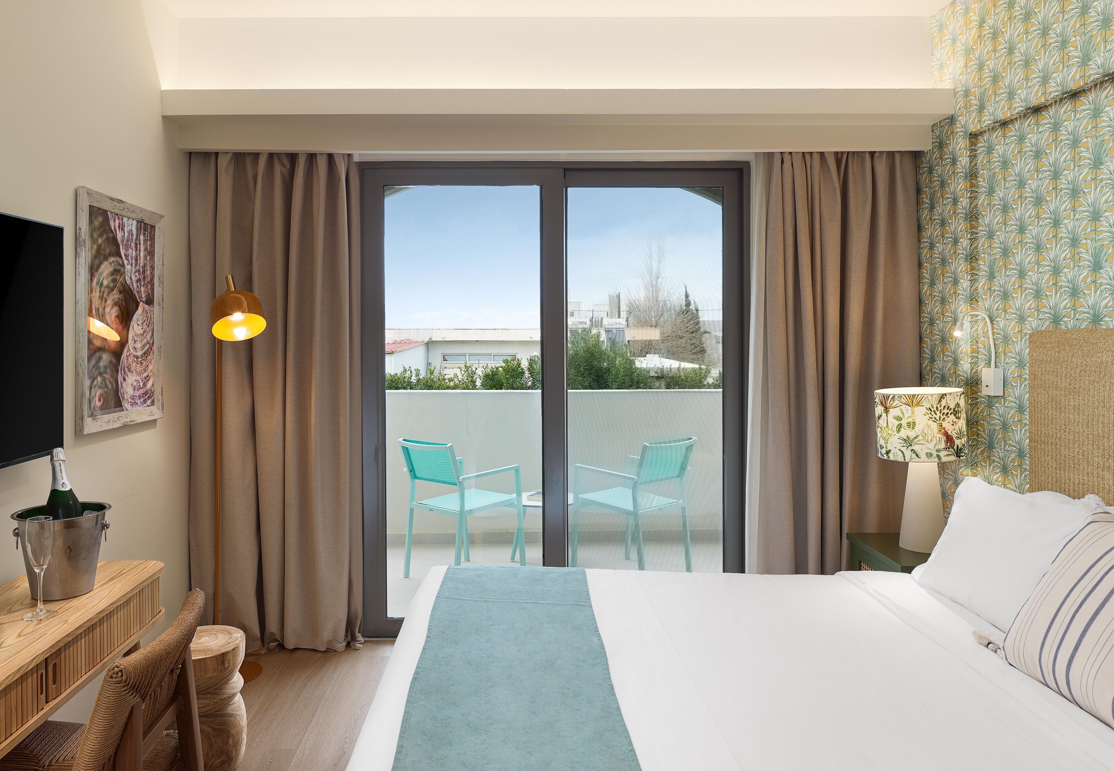 A hotel room with a bed, a desk with a bottle and glass, two chairs, a balcony with a view, and a TV on the wall.