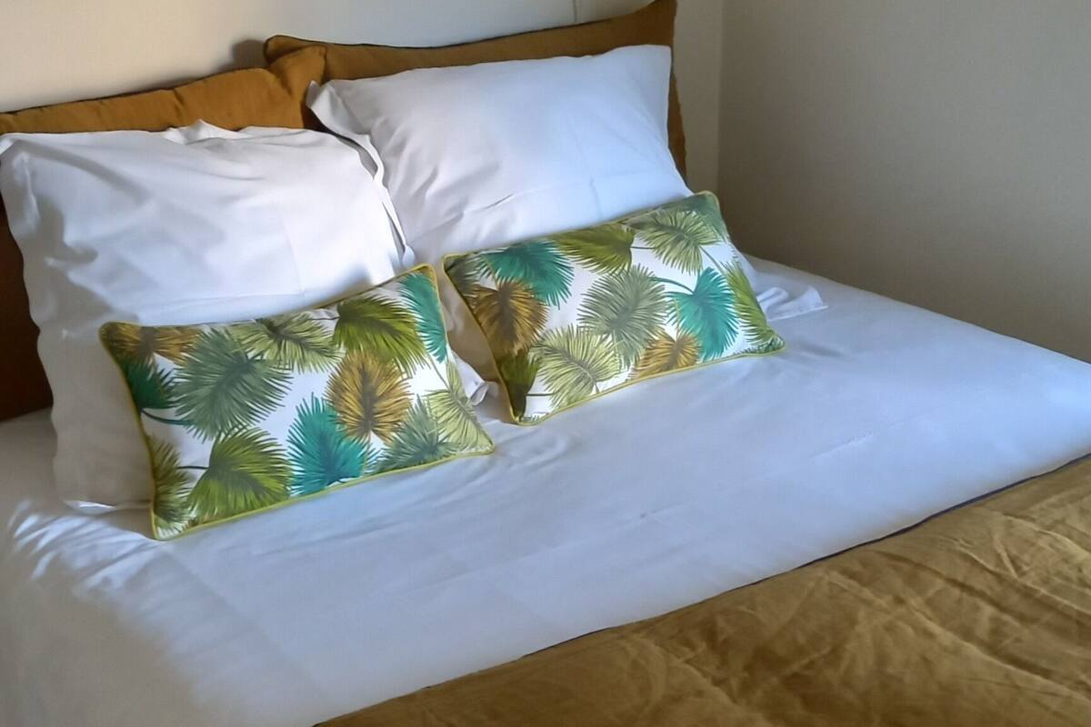 Un lit bien fait, avec une literie blanche et marron, un coussin orné d’un motif de palmiers tropicaux et une simple lampe blanche posée à côté.