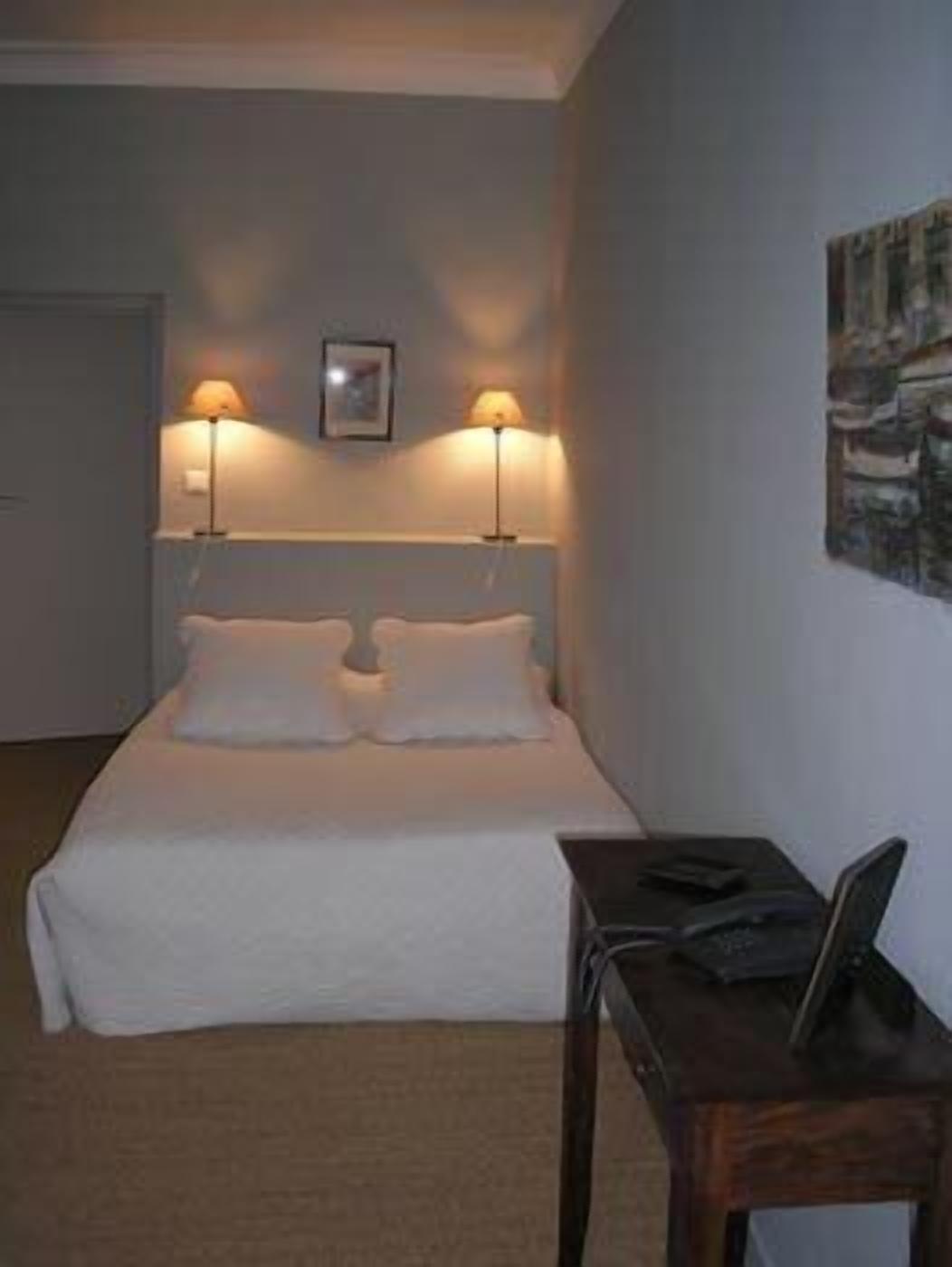Quarto de hotel com uma cama, duas luminárias de cabeceira, uma pequena mesa com um laptop e um quadro na parede.