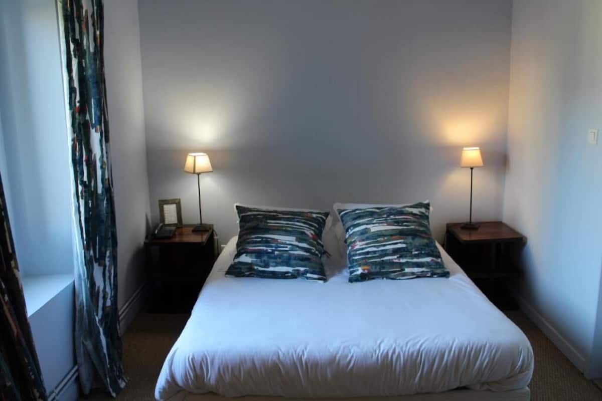 Un lit simple avec une literie blanche et deux oreillers assortis, flanqué de deux tables de chevet avec des lampes, dans une pièce aux murs unis et ornée d’un rideau sur un des côtés.
