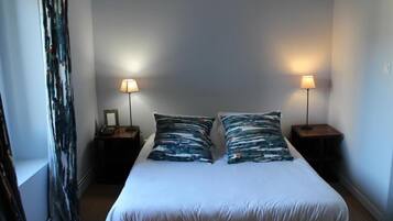 Een eenpersoonsbed met wit beddengoed en twee bijpassende kussens, met aan weerszijden nachtkastjes met lampen, in een kamer met eenvoudige wanden en een gordijn aan één kant.
