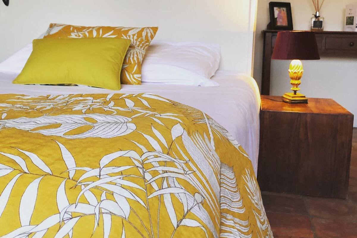 Une chambre d’hôtel avec un lit recouvert d’une courtepointe à motifs floraux jaune et blanc, un oreiller vert et un oreiller blanc. On y trouve une table de chevet avec une lampe et une petite étagère ornée d’objets décoratifs.