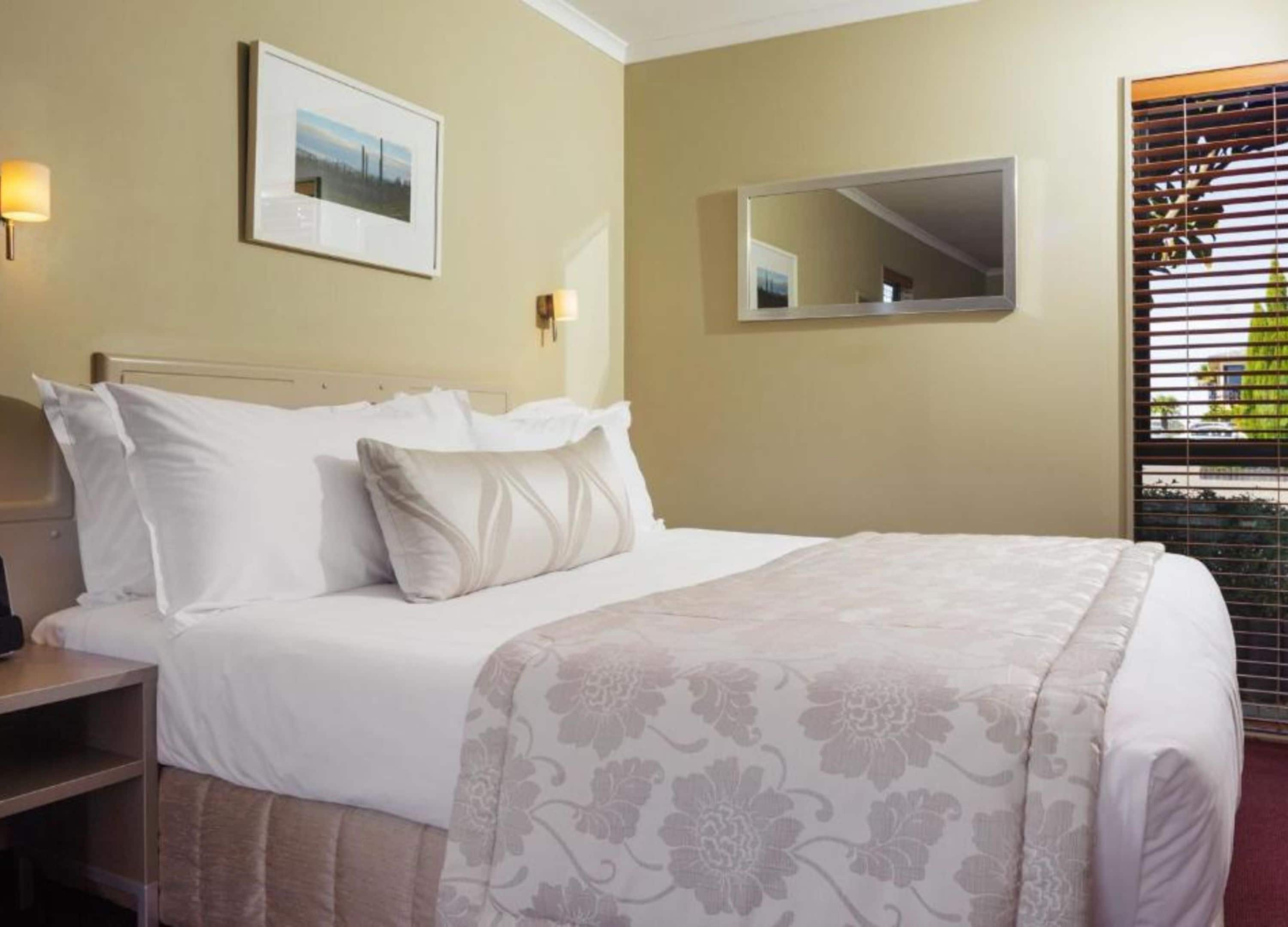 A neatly made bed with white linens and a floral-patterned blanket, a bedside table with a lamp, a framed picture on the wall, and a window with blinds.