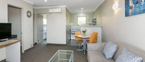 A compact living space with a sofa, a glass coffee table, a TV, a dining area with chairs, and a kitchen in the background.
