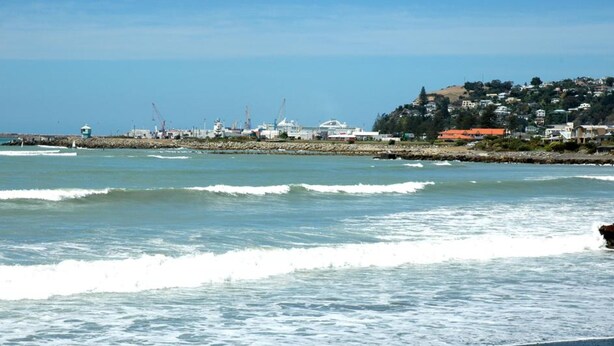Paisaje costero con olas, una costa rocosa y edificios al fondo.