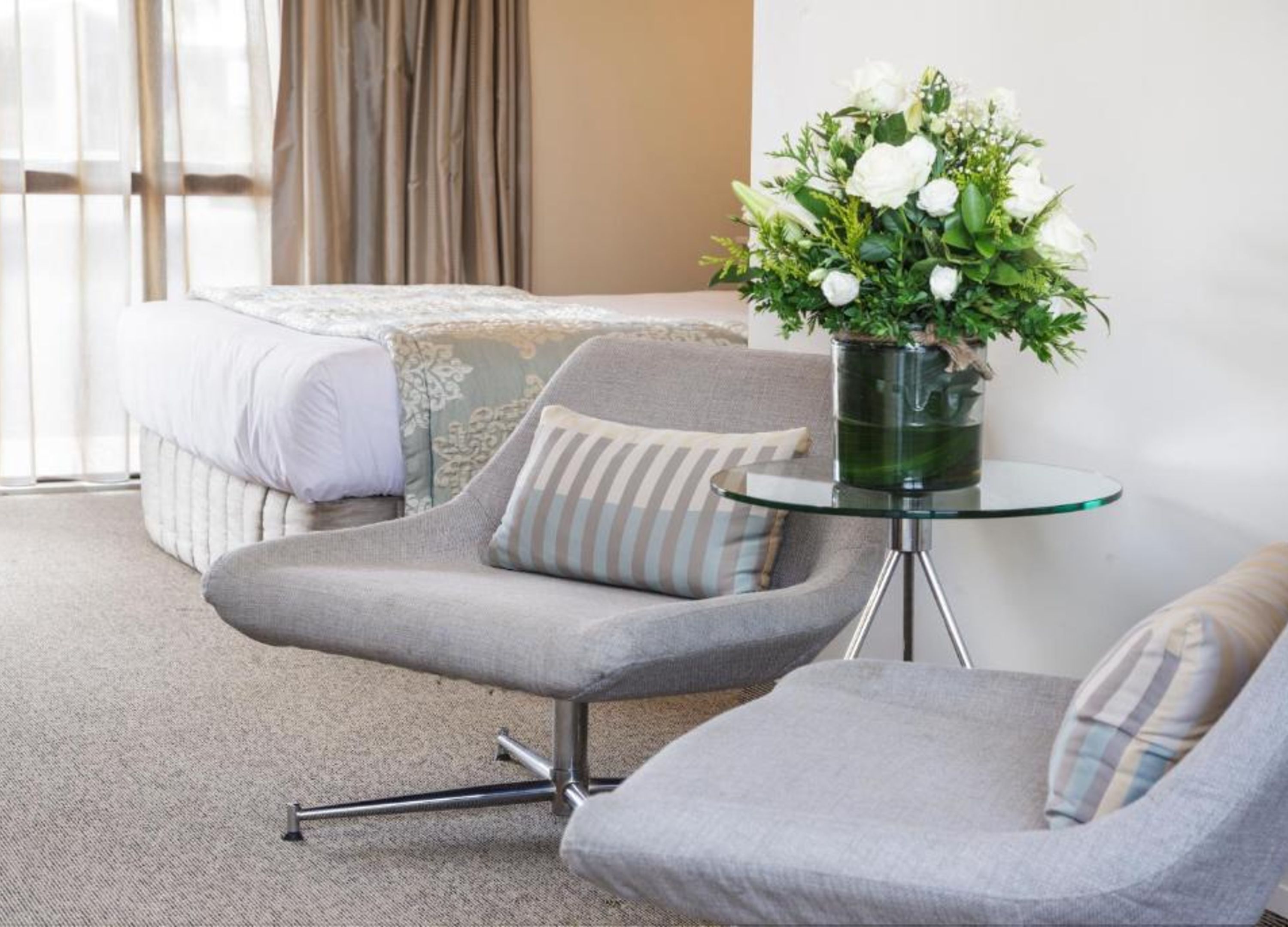 Habitación de hotel con una cama, dos sillones grises, una mesita de vidrio con un florero de flores blancas y vistas al exterior a través de cortinas traslúcidas.