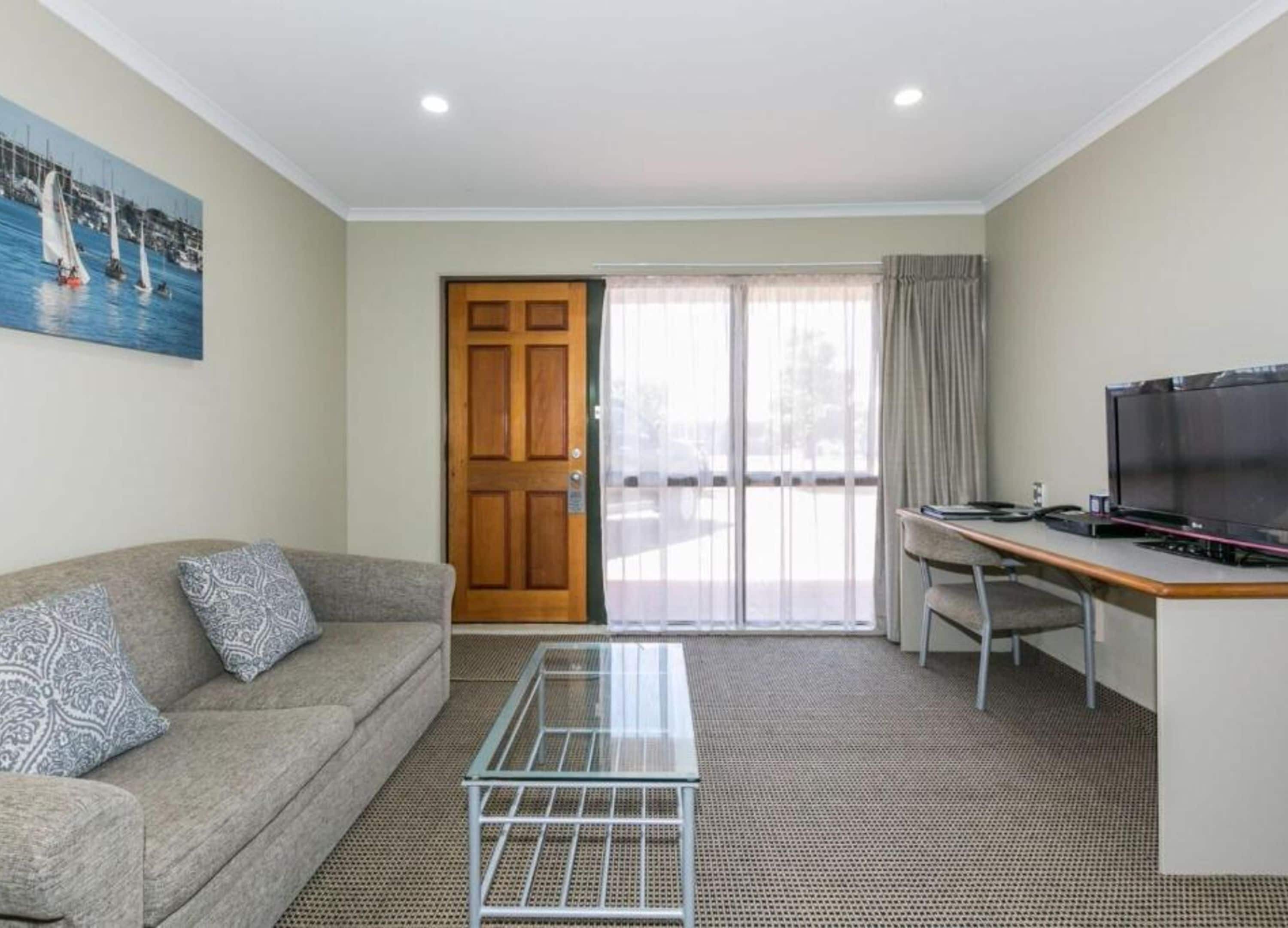 A hotel room with a sofa, a glass coffee table, a desk with a computer, a television, and a door leading to a balcony.