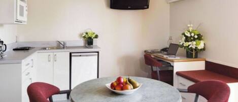 Une pièce compacte avec un coin cuisine, une table ronde sur laquelle se trouve un saladier de fruits, deux chaises rouges, un téléviseur fixé au mur et un bureau avec un ordinateur portable et des fleurs.