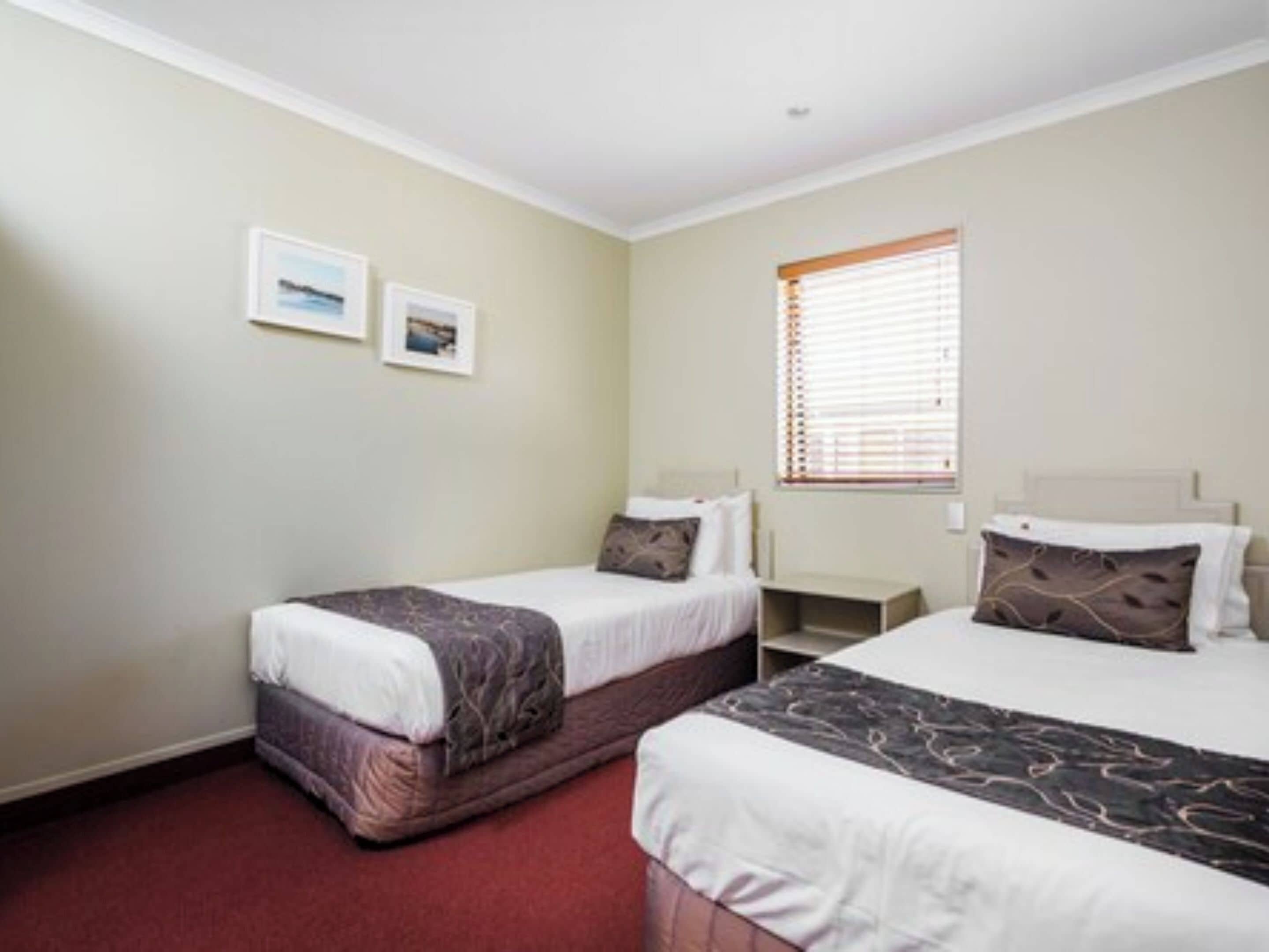 A hotel room with two single beds, a small bedside table, a nightstand, and a window with blinds.