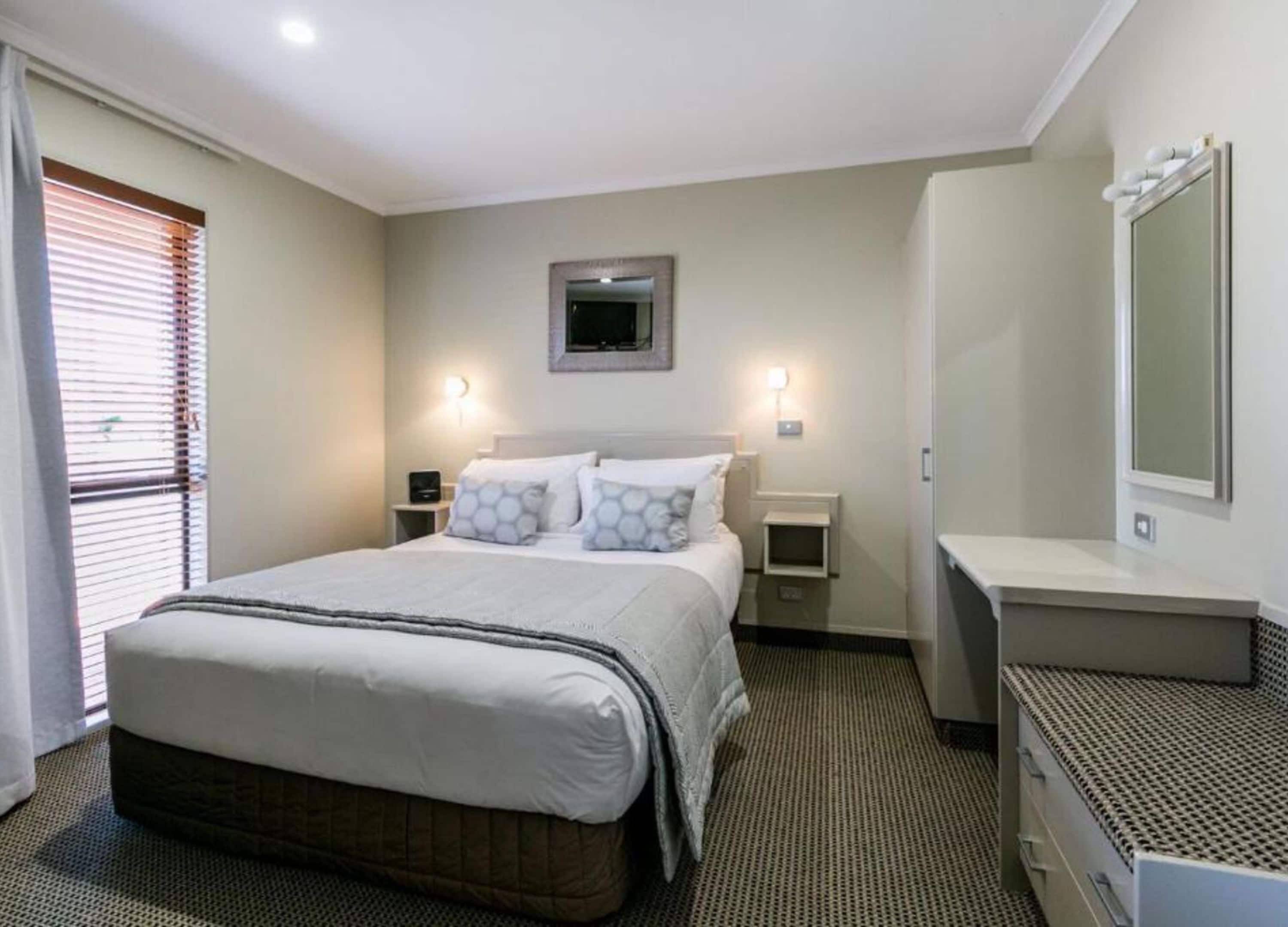 Una habitación de hotel con una cama grande, mesitas de noche, un espejo y una ventana con persianas.