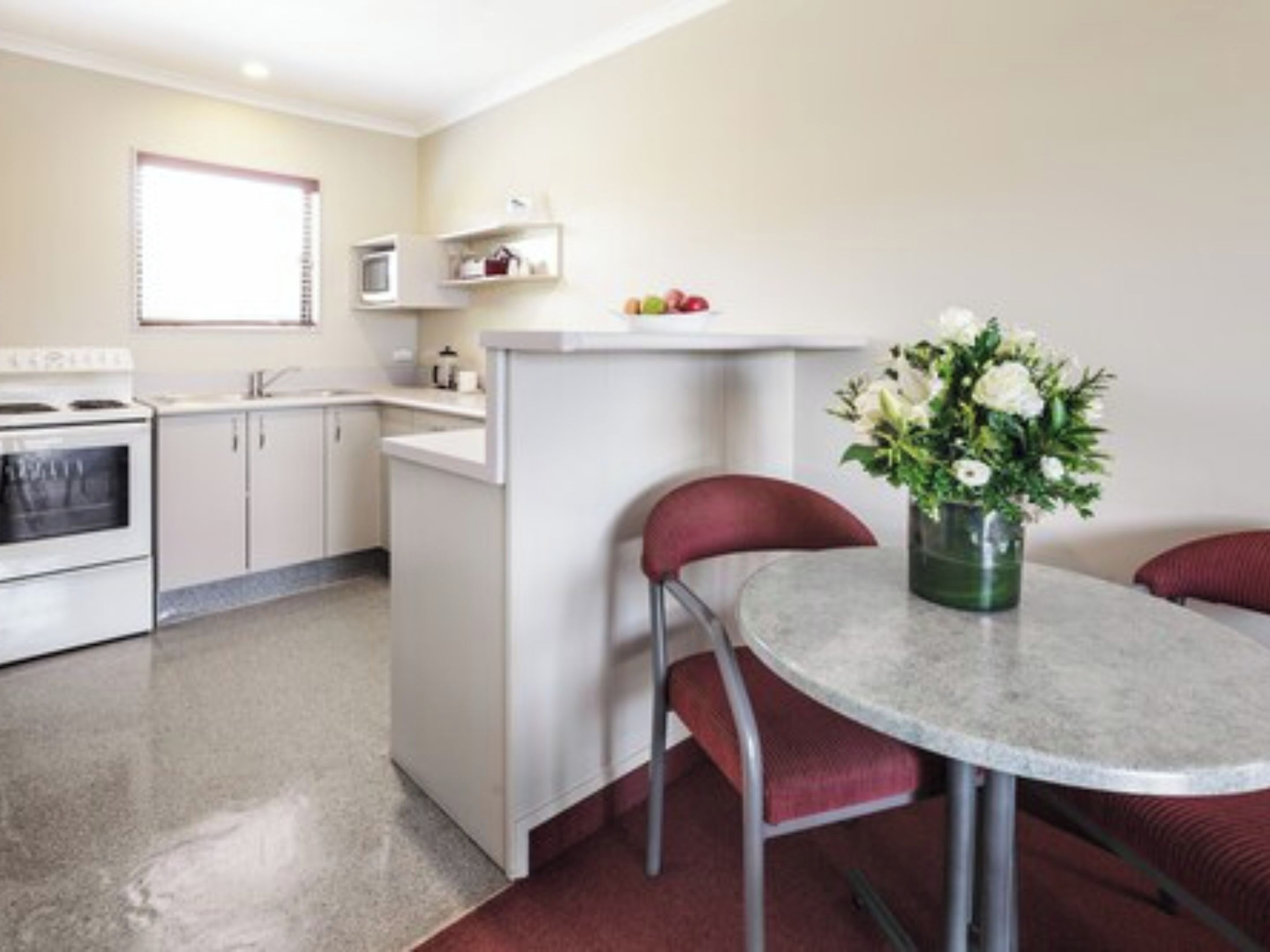 A compact kitchen with white cabinets, a stove, and a microwave. There's a small dining area with a table and two chairs. A vase with flowers is on the table.