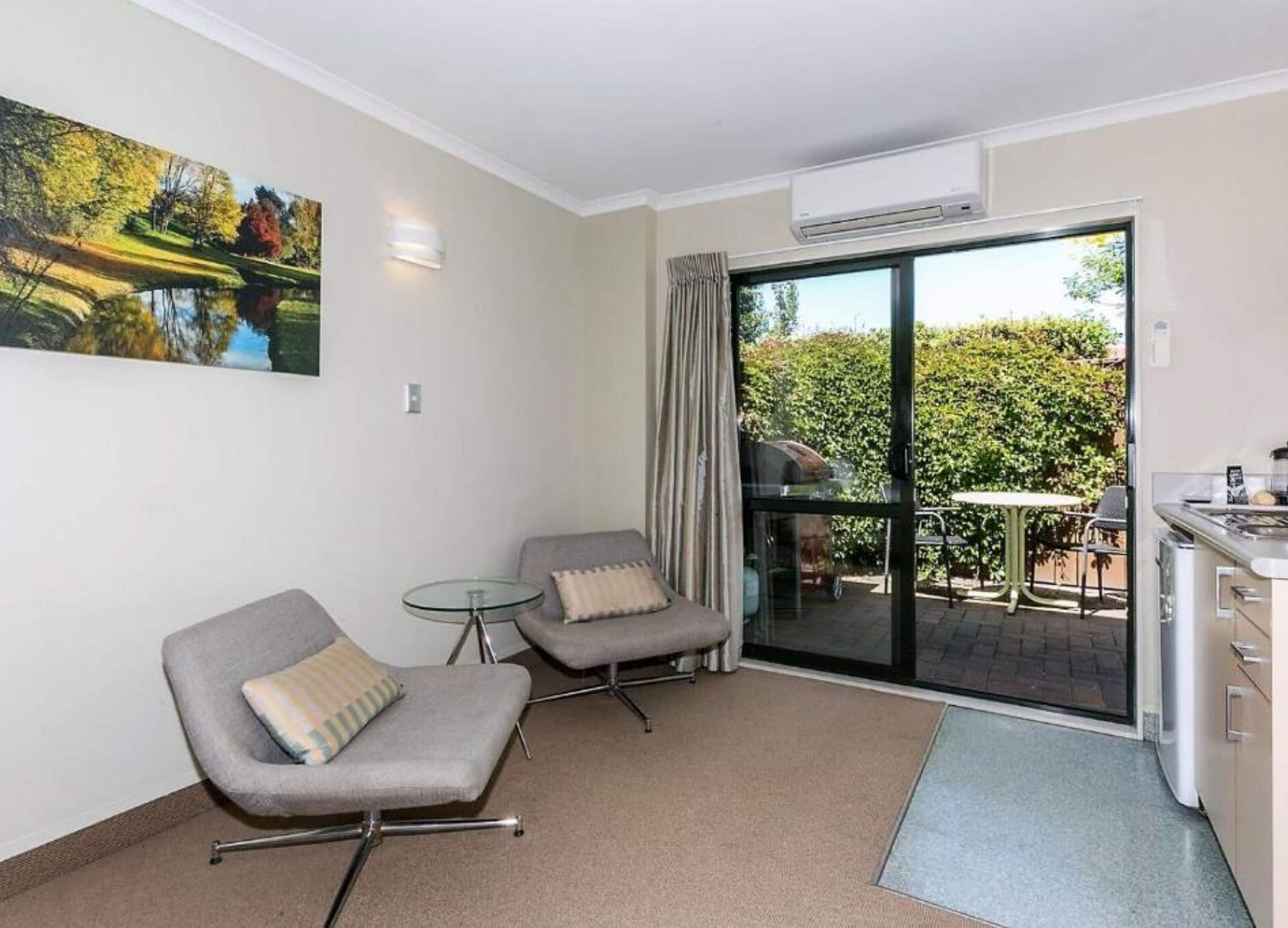 A modern room with two grey armchairs, a glass table, a painting of a landscape, a sliding glass door to an outdoor area with a barbecue, and a kitchenette.