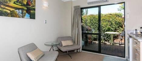A modern room with two grey armchairs, a glass table, a painting of a landscape, a sliding glass door to an outdoor area with a barbecue, and a kitchenette.