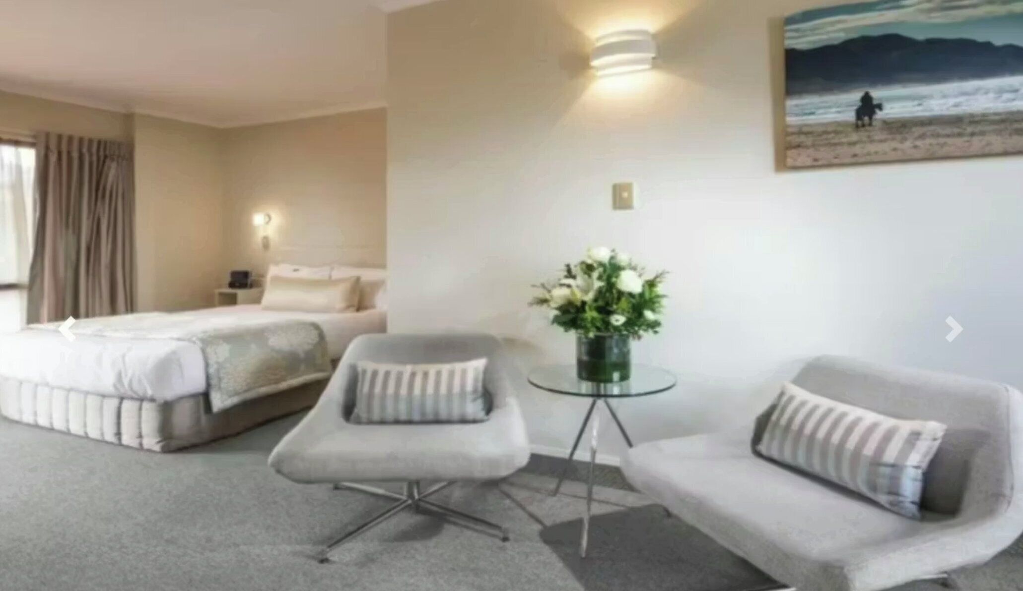 Habitación de hotel con una cama, dos sillones, una mesita con un florero y un cuadro de una playa en la pared.