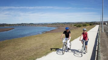 Two cyclists on a paved path beside a river with a bridge in the background.