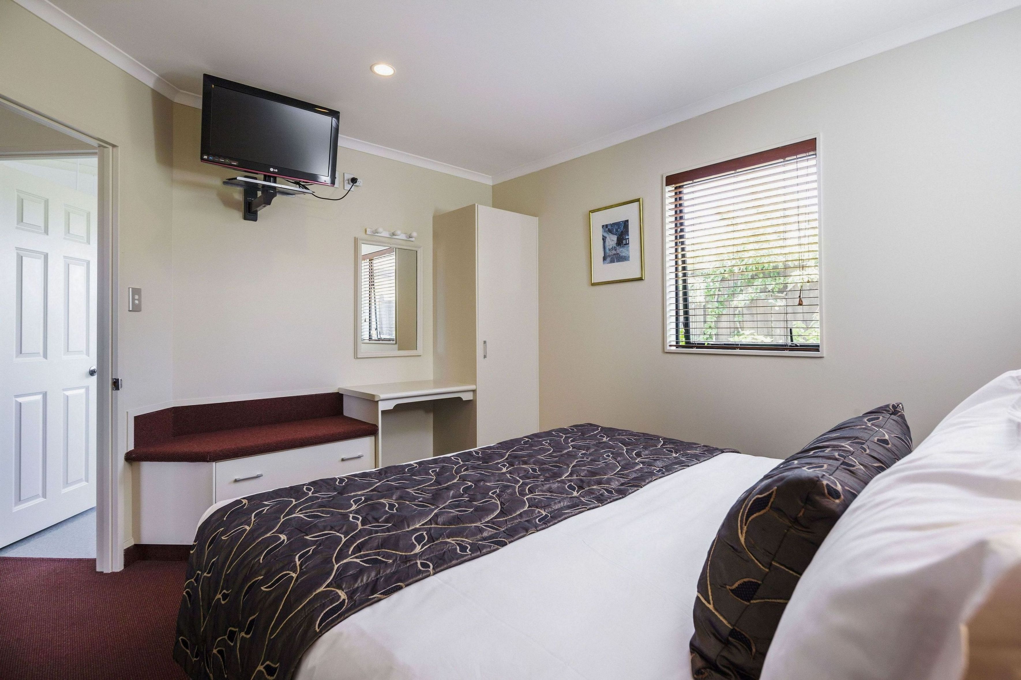 Habitación de hotel con cama, escritorio, televisor montado en la pared, ventana con persianas y puerta.
