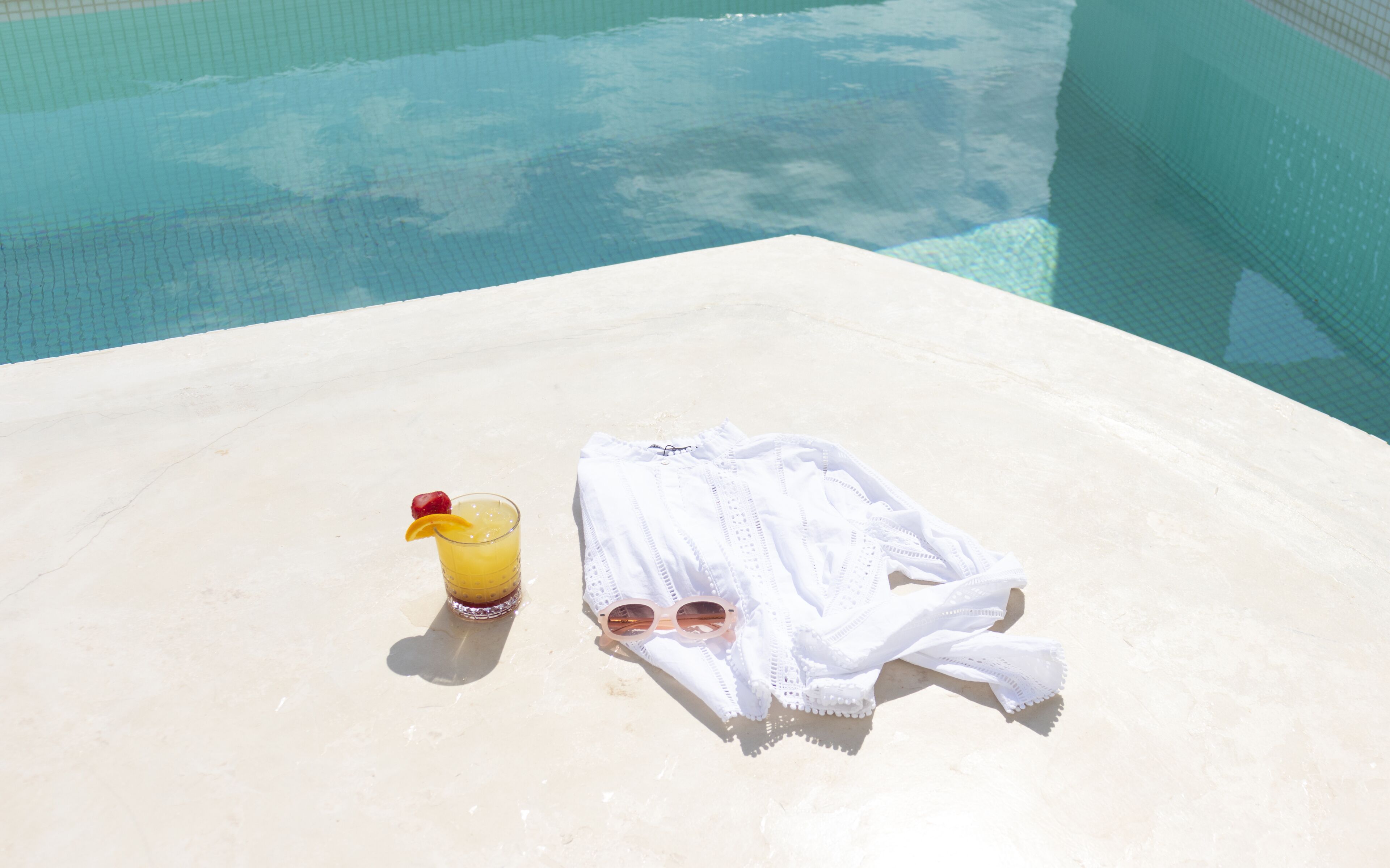 Cenário à beira da piscina com toalha, óculos de sol e uma bebida sobre uma superfície branca.