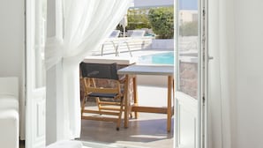 A bedroom with a bed, a table, and a chair, leading to a balcony with a pool.