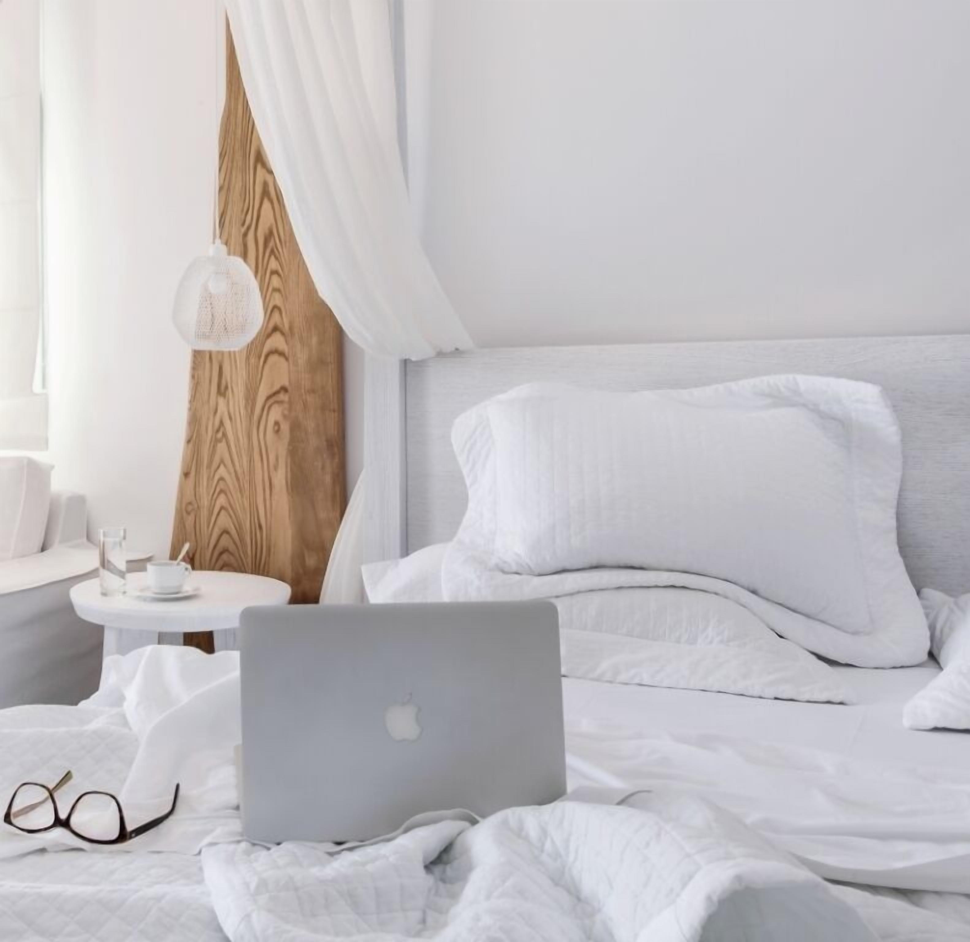 A neatly made bed with white bedding, a laptop, and a pair of glasses on the bed.