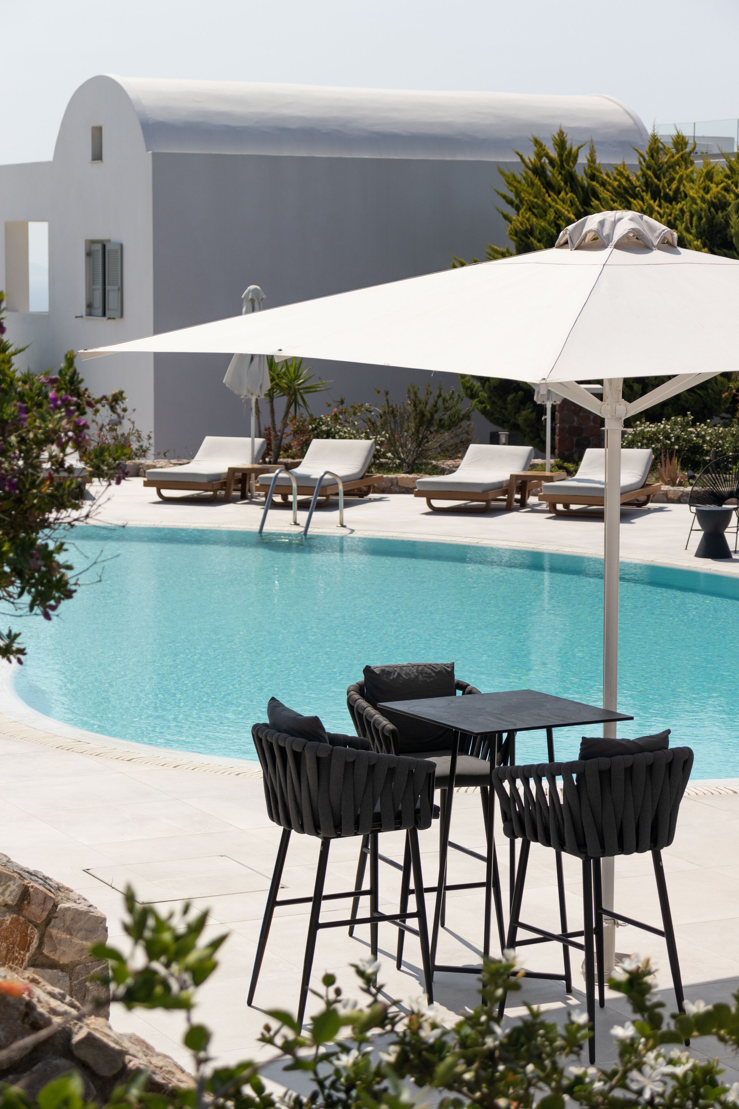 A pool area with lounge chairs and a table, surrounded by a white building and greenery.