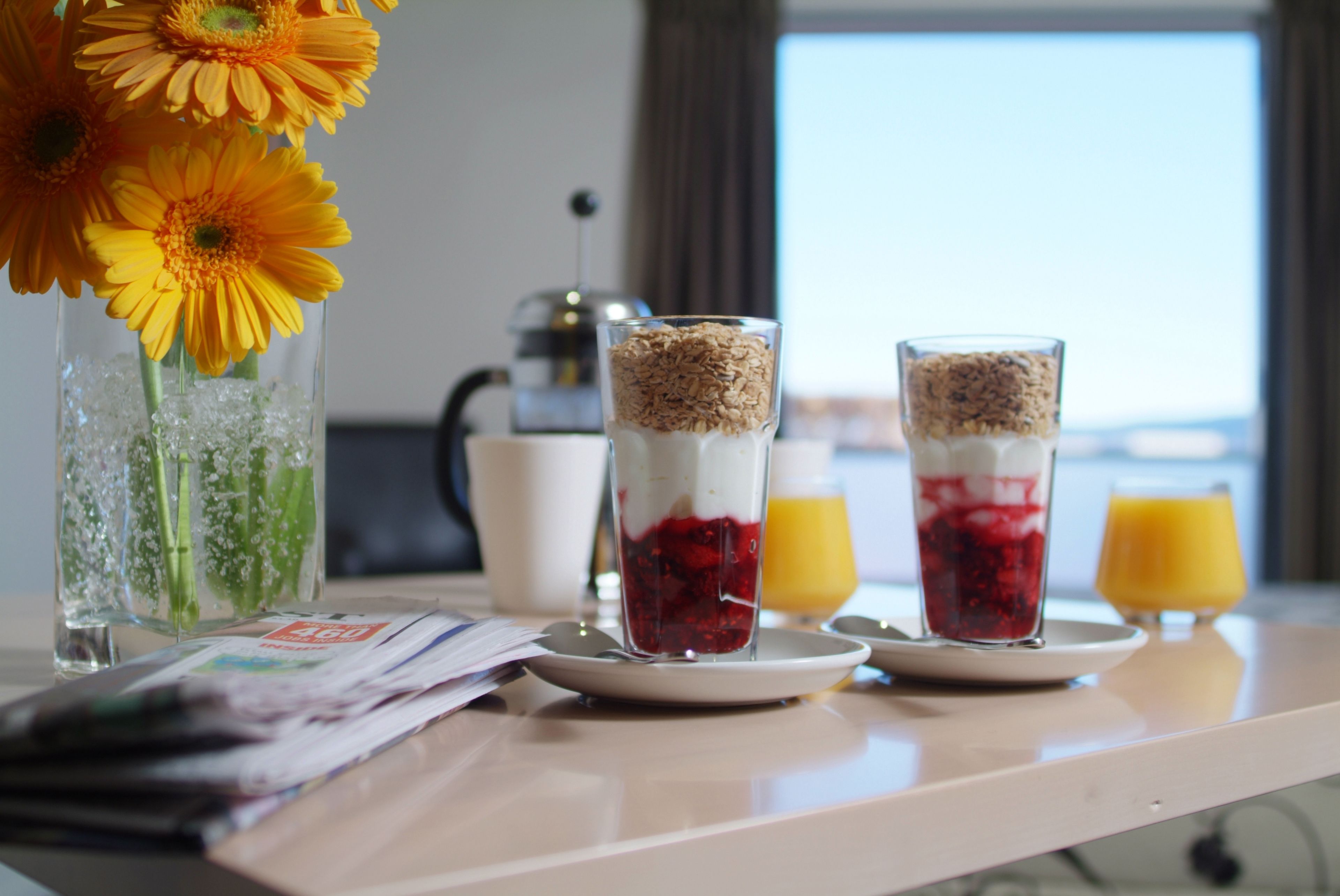 Une table de petit-déjeuner avec un vase de fleurs jaunes, deux verres contenant des couches de fruits et de céréales, et une machine à café en arrière-plan.