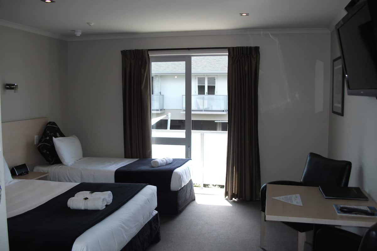 A hotel room with two beds, a desk, and a chair. There is a window with curtains and a view of a building outside.