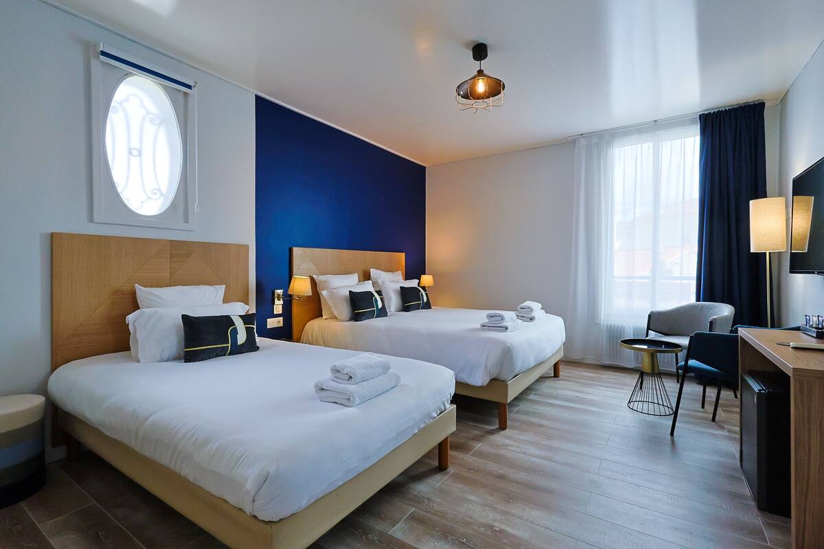 A hotel room with two beds, a desk, a chair, a TV, and a window with curtains.