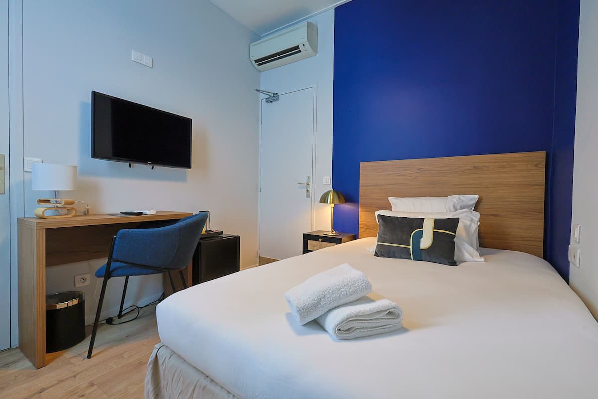 A hotel room with a bed, a desk, a chair, a TV, and an air conditioning unit.