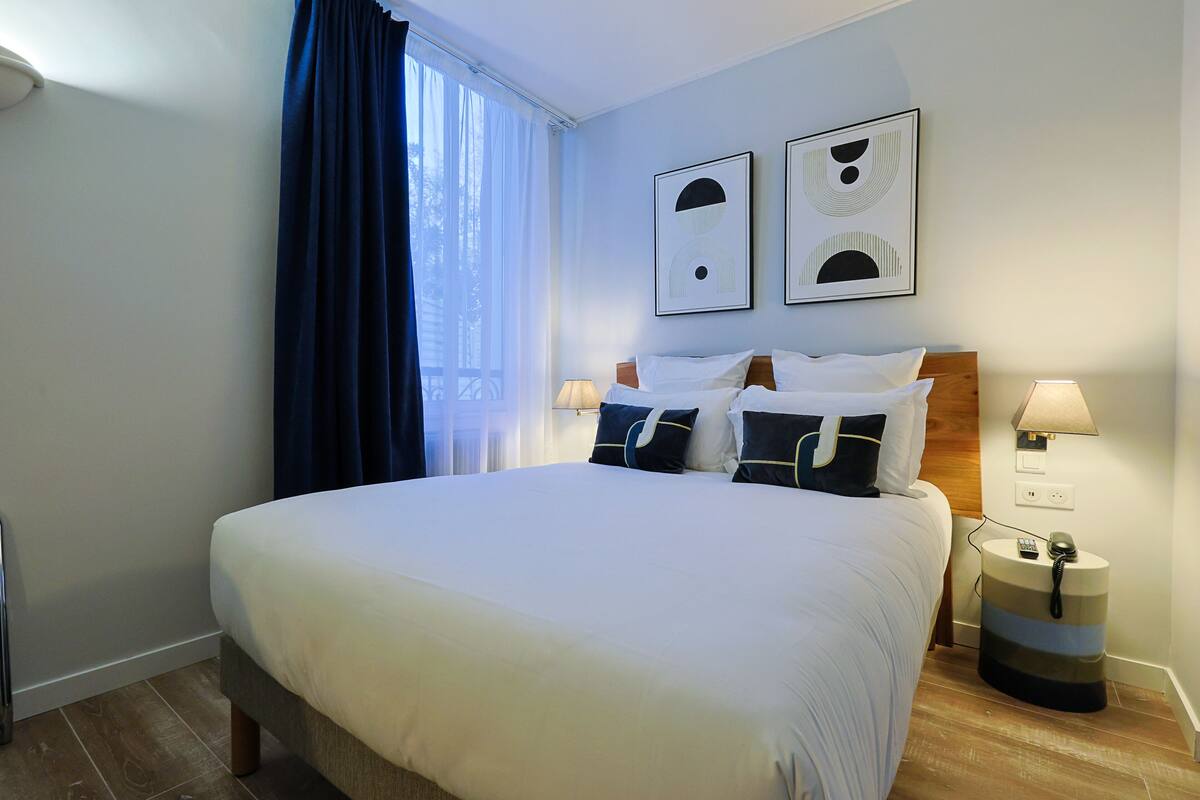 A neatly made bed with white linens and dark pillows, a bedside lamp, and two framed abstract artworks on the wall.