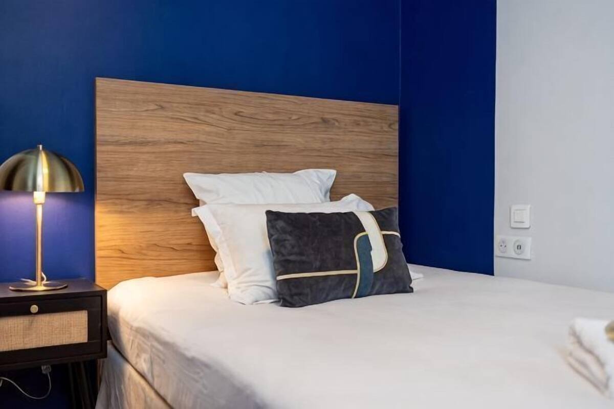 A neatly made bed with white and dark pillows, a wooden headboard, a bedside lamp, and a nightstand with a lamp.