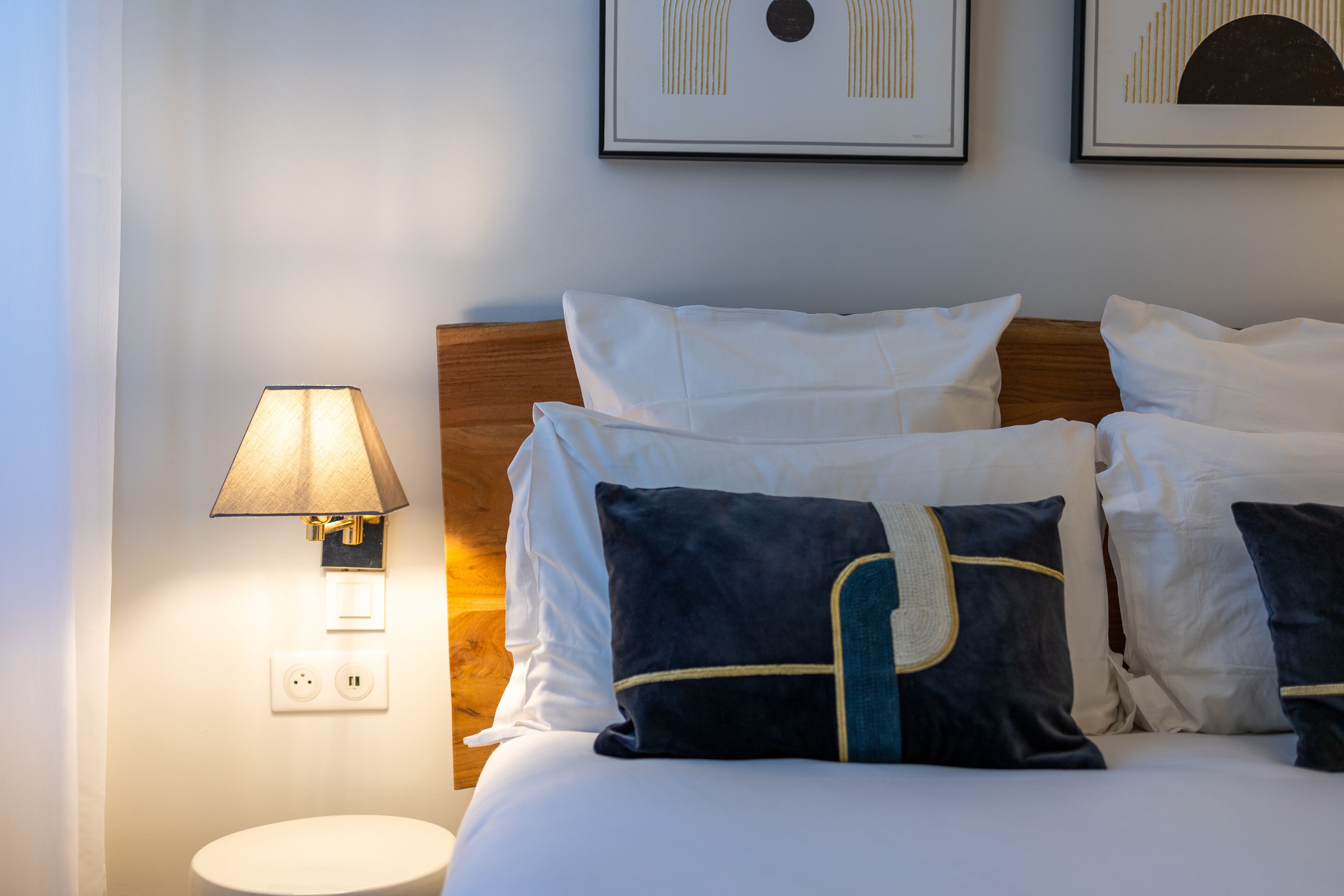 Una cama bien tendida con almohadas blancas y azul oscuro, un cabecero de madera y una lámpara de pared.