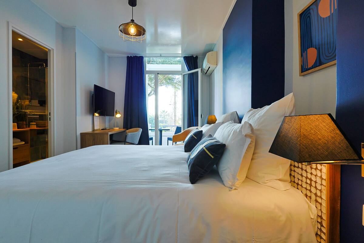 A hotel room with a large bed, a desk, and a chair. There is a TV on the wall and a window with curtains.