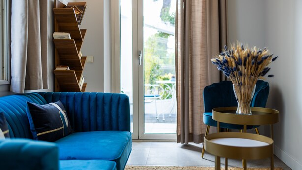 A modern living room with a blue sofa, a wooden bookshelf, a blue armchair, and a small round table with a vase of dried flowers. - Hotel Albert 1er (Cannes)