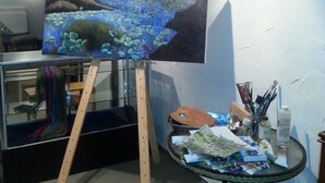 An art studio with an easel holding a landscape painting, a small table with art supplies, and a painting of tulips on the wall.
